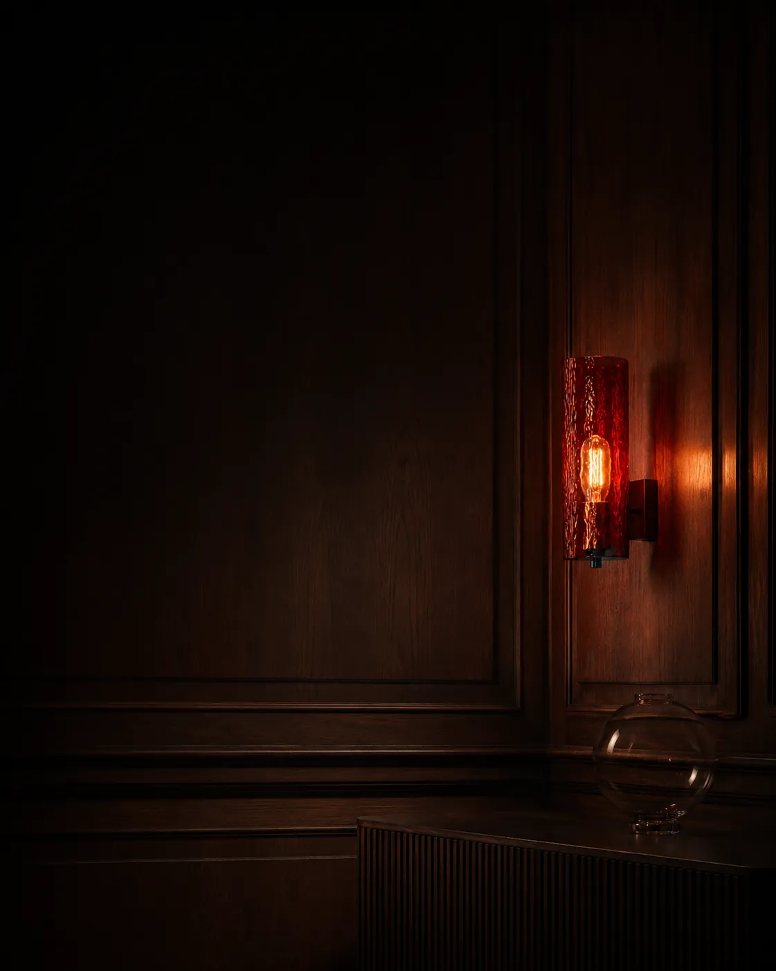A dark panelled room at night lit only by a red glass wall sconce, with a small lit tree and glassware catching the glow at the far end